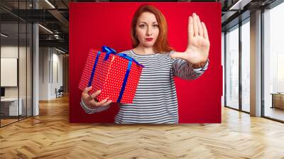 Young beautiful redhead woman holding birthday gift over red isolated background with open hand doing stop sign with serious and confident expression, defense gesture Wall mural