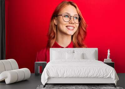 Young beautiful redhead woman wearing casual clothes and glasses over red background looking away to side with smile on face, natural expression. laughing confident. Wall mural