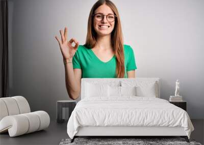 Young beautiful redhead woman wearing casual green t-shirt and glasses over white background smiling positive doing ok sign with hand and fingers. Successful expression. Wall mural