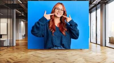 Young beautiful redhead woman wearing casual sweater and glasses over blue background smiling cheerful showing and pointing with fingers teeth and mouth. dental health concept. Wall mural