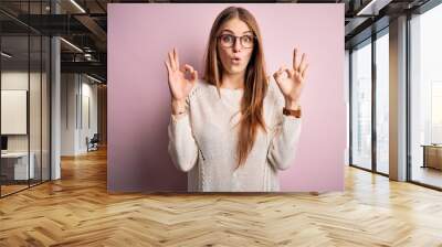 Young beautiful redhead woman wearing casual sweater and glasses over pink background looking surprised and shocked doing ok approval symbol with fingers. Crazy expression Wall mural