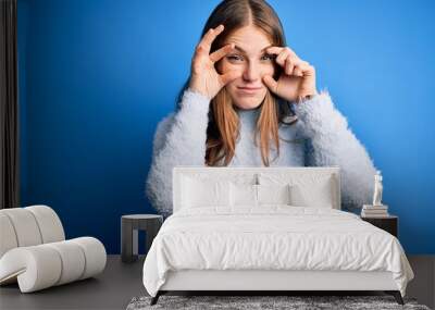 Young beautiful redhead woman wearing casual sweater over isolated blue background Trying to open eyes with fingers, sleepy and tired for morning fatigue Wall mural