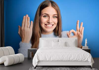 Young beautiful redhead woman wearing wedding ring on finger over blue background doing ok sign with fingers, excellent symbol Wall mural