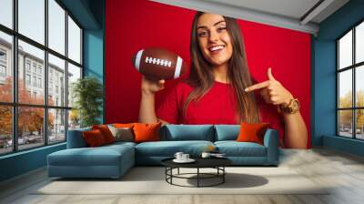 Young beautiful sportswoman holding football ball over isolated red background with surprise face pointing finger to himself Wall mural