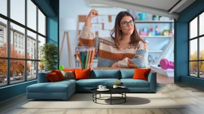 Young beautiful teacher woman wearing sweater and glasses sitting on desk at kindergarten Strong person showing arm muscle, confident and proud of power Wall mural