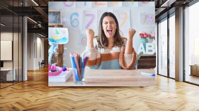 Young beautiful teacher woman wearing sweater and glasses sitting on desk at kindergarten very happy and excited doing winner gesture with arms raised, smiling and screaming for success.  Wall mural