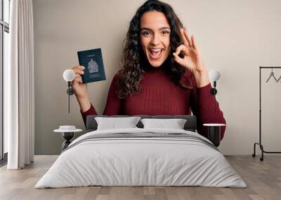 Young beautiful tourist woman with curly hair on vacation holding canadian canada passport doing ok sign with fingers, excellent symbol Wall mural