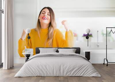 Young beautiful woman drinking a cup of coffee at home very happy and excited doing winner gesture with arms raised, smiling and screaming for success. Celebration concept. Wall mural