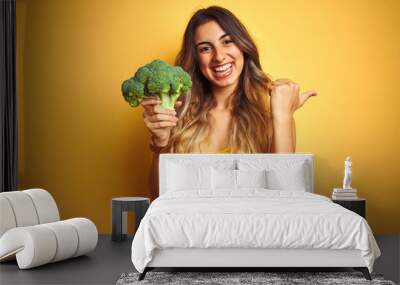 Young beautiful woman eating broccoli over yellow isolated background pointing and showing with thumb up to the side with happy face smiling Wall mural