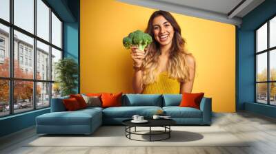 Young beautiful woman eating broccoli over yellow isolated background with a happy face standing and smiling with a confident smile showing teeth Wall mural