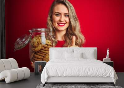 Young beautiful woman holding a jar of cookies over red isolated background happy with big smile doing ok sign, thumb up with fingers, excellent sign Wall mural