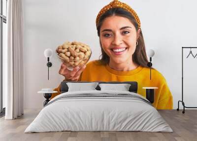 Young beautiful woman holding bowl with peanuts standing over isolated white background with a happy face standing and smiling with a confident smile showing teeth Wall mural