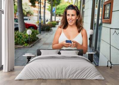 Young beautiful woman smiling happy walking on city streets on a sunny day of summer using smartphone Wall mural