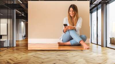 Young beautiful woman smiling sitting on the floor and looking at the phone. Wall mural