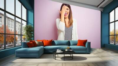 Young beautiful woman standing over pink isolated background covering one eye with hand, confident smile on face and surprise emotion. Wall mural