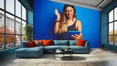 Young beautiful woman using touchpad tablet over blue isolated background very happy and excited, winner expression celebrating victory screaming with big smile and raised hands Wall mural