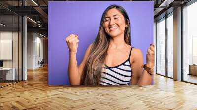 Young beautiful woman wearing casual summer striped top over purple isolated background very happy and excited doing winner gesture with arms raised, smiling and screaming for success. Celebration Wall mural