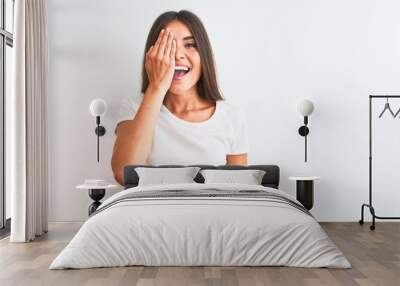 Young beautiful woman wearing casual t-shirt standing over isolated white background covering one eye with hand, confident smile on face and surprise emotion. Wall mural