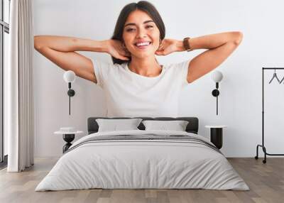 Young beautiful woman wearing casual t-shirt standing over isolated white background relaxing and stretching, arms and hands behind head and neck smiling happy Wall mural