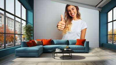 Young beautiful woman wearing casual white t-shirt over isolated background doing happy thumbs up gesture with hand. Approving expression looking at the camera showing success. Wall mural