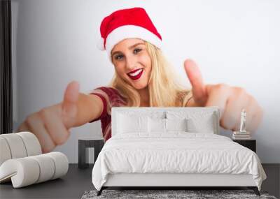 Young beautiful woman wearing Christmas Santa hat over isolated white background approving doing positive gesture with hand, thumbs up smiling and happy for success. Winner gesture. Wall mural