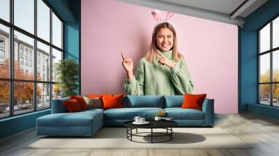 Young beautiful woman wearing easter rabbit ears standing over isolated pink background smiling and looking at the camera pointing with two hands and fingers to the side. Wall mural