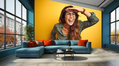 Young beautiful woman wearing green shirt and glasses over yelllow isolated background very happy and smiling looking far away with hand over head. Searching concept. Wall mural