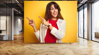 Young beautiful woman wearing red t-shirt and stripes shirt over yellow isolated background smiling and looking at the camera pointing with two hands and fingers to the side. Wall mural