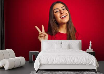 Young beautiful woman wearing t-shirt standing over isolated red background smiling with happy face winking at the camera doing victory sign. Number two. Wall mural