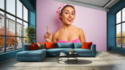 Young beautiful woman wearing towel and shower cap over isolated pink background pointing finger up with successful idea. Exited and happy. Number one. Wall mural