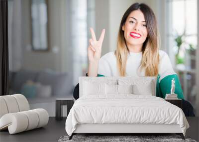 Young beautiful woman wearing winter sweater at home showing and pointing up with fingers number two while smiling confident and happy. Wall mural