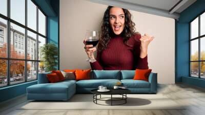 Young beautiful woman with curly hair drinking glass of red wine over white background pointing and showing with thumb up to the side with happy face smiling Wall mural