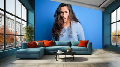 Young beautiful woman with curly hair wearing blue casual sweater over isolated background In shock face, looking skeptical and sarcastic, surprised with open mouth Wall mural