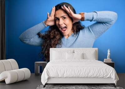 Young beautiful woman with curly hair wearing blue casual sweater over isolated background Smiling cheerful playing peek a boo with hands showing face. Surprised and exited Wall mural