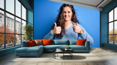 Young beautiful woman with curly hair wearing blue casual sweater over isolated background success sign doing positive gesture with hand, thumbs up smiling and happy. Cheerful expression and winner Wall mural