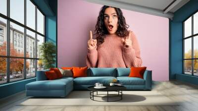 Young beautiful woman with curly hair wearing casual sweater over isolated pink background amazed and surprised looking up and pointing with fingers and raised arms. Wall mural