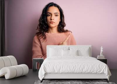 Young beautiful woman with curly hair wearing casual sweater over isolated pink background Pointing with hand finger to the side showing advertisement, serious and calm face Wall mural