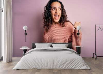 Young beautiful woman with curly hair wearing casual sweater over isolated pink background smiling with happy face looking and pointing to the side with thumb up. Wall mural