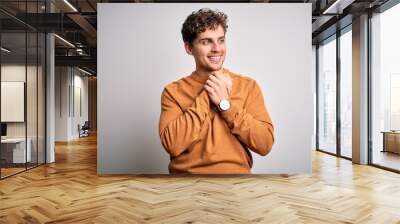 Young blond handsome man with curly hair wearing casual sweater over white background laughing nervous and excited with hands on chin looking to the side Wall mural