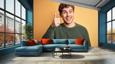 Young blond handsome man with curly hair wearing green sweater over yellow background doing happy thumbs up gesture with hand. Approving expression looking at the camera showing success. Wall mural