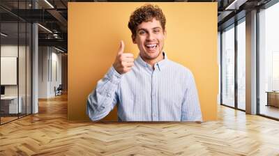 Young blond handsome man with curly hair wearing striped shirt over yellow background doing happy thumbs up gesture with hand. Approving expression looking at the camera showing success. Wall mural