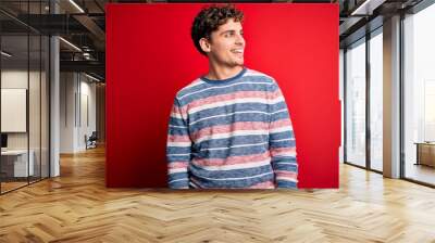 Young blond handsome man with curly hair wearing striped sweater over red background looking away to side with smile on face, natural expression. Laughing confident. Wall mural