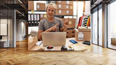 Young blond man wearing operator headset working at online shop smiling and looking at the camera pointing with two hands and fingers to the side. Wall mural