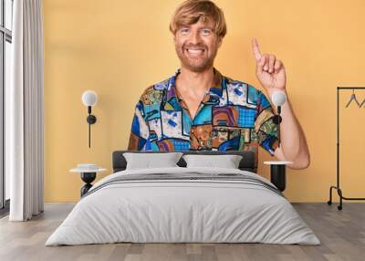 Young blond man wearing summer shirt showing and pointing up with finger number one while smiling confident and happy. Wall mural