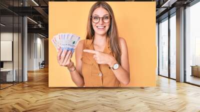 Young blonde girl holding colombian pesos smiling happy pointing with hand and finger Wall mural