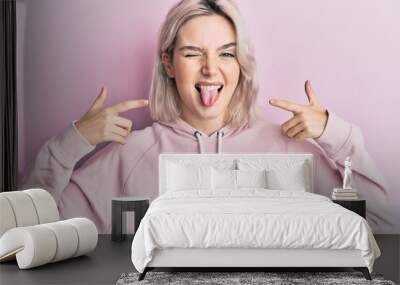 Young blonde girl pointing mouth with fingers sticking tongue out happy with funny expression. Wall mural