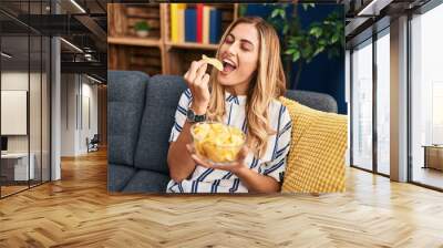 Young blonde woman eating chips potatoes sitting on sofa at home Wall mural