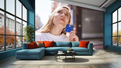 Young blonde woman holding credit card with doubt expression at street Wall mural