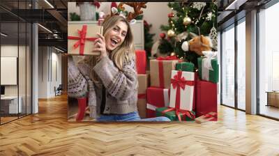 Young blonde woman holding gift sitting by christmas tree at home Wall mural