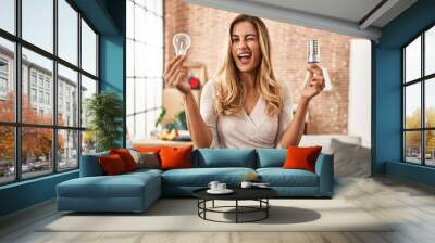 Young blonde woman holding led lightbulb and incandescent bulb winking looking at the camera with sexy expression, cheerful and happy face. Wall mural